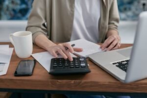 Small business owner using a calculator and laptop to review finances at a wooden desk