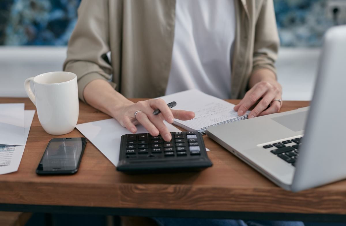 Small business owner using a calculator and laptop to review finances at a wooden desk