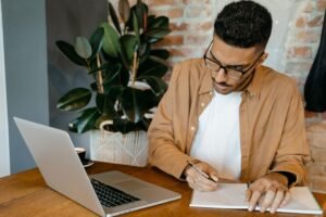 Man writing notes at a desk with a laptop open beside him.