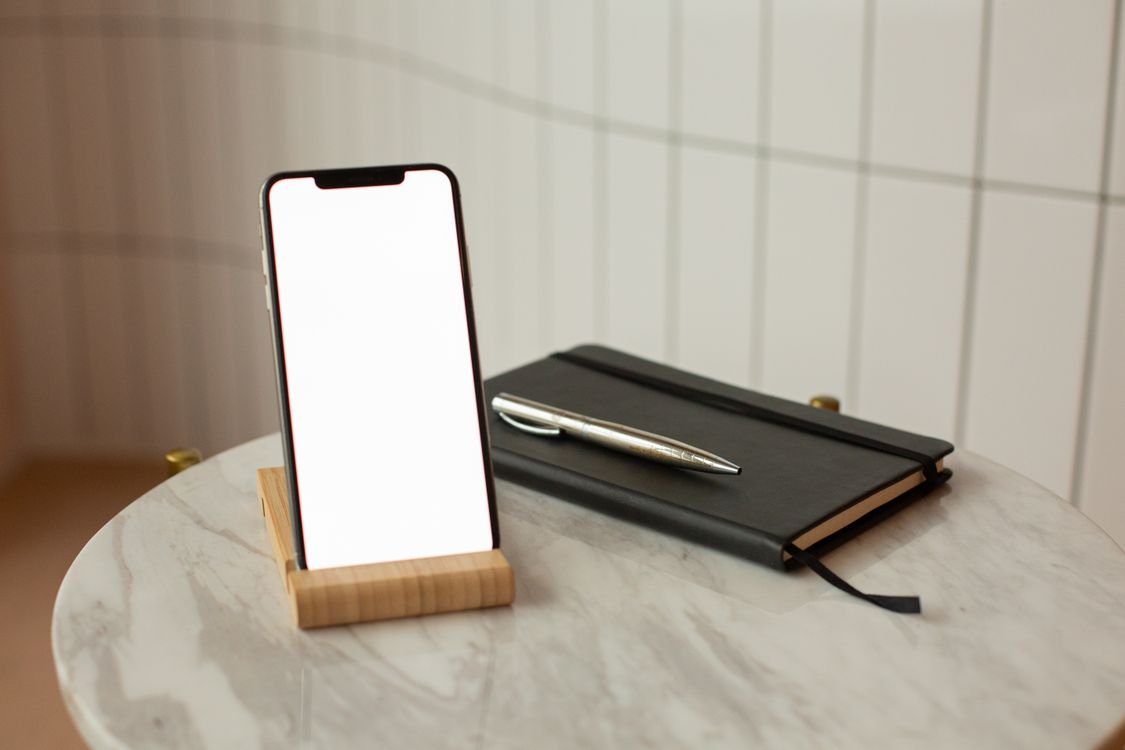 Smartphone with blank screen resting on a wooden stand beside a notebook and pen