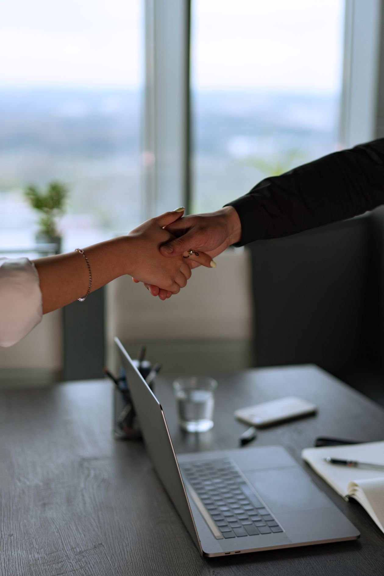 Two business people shaking hands across a desk with a laptop in a modern office