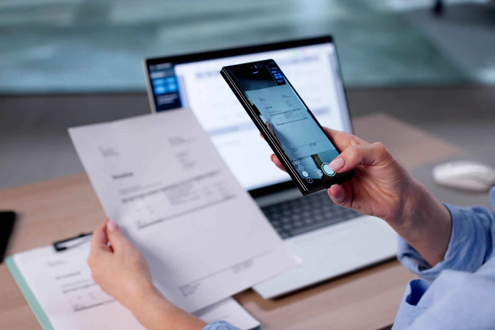 Small business owner photographing a paper invoice with a smartphone, laptop open in the background