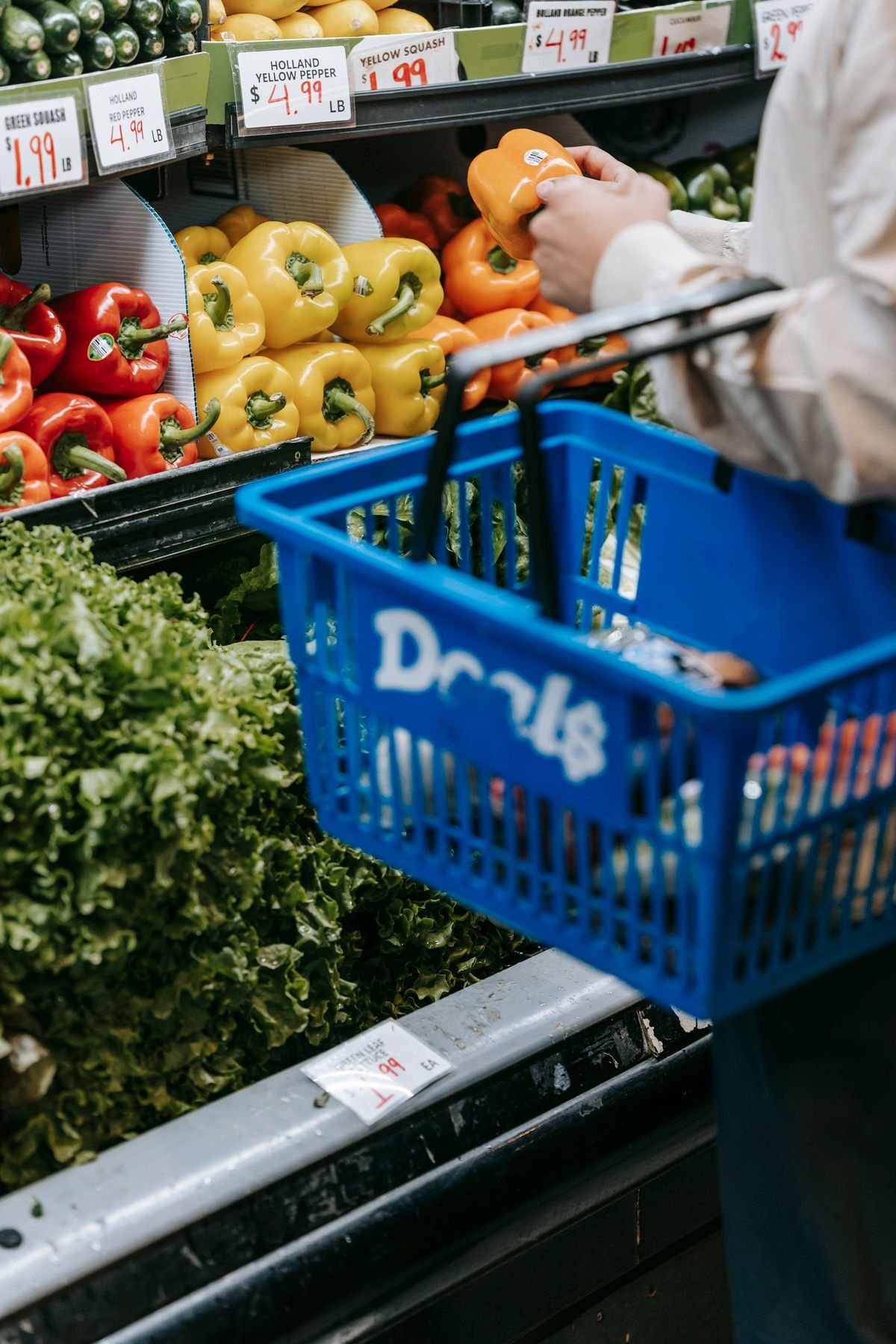 Shopper holding an orange bell pepper above a blue shopping basket in a grocery store
