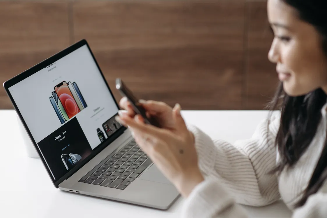 Woman checking product prices on a laptop while holding a phone