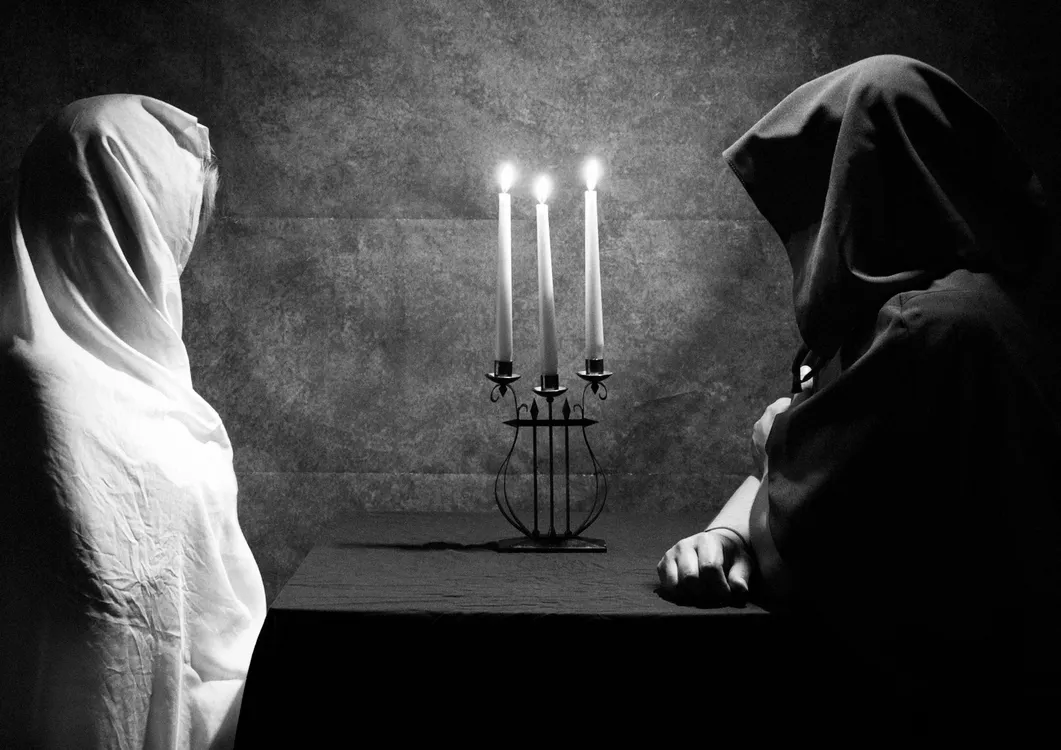 Two figures in cloaks seated beside candles in a dark round table scene