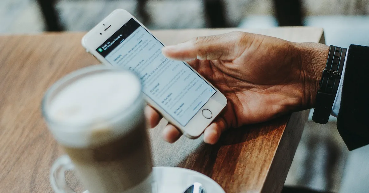 Hand scrolling through an email inbox on a smartphone beside a cup of coffee